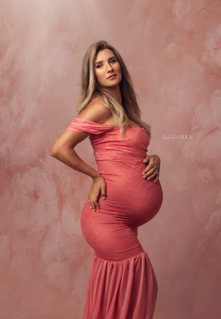 gorgeous pregnant woman in coral colored couture maternity gown in photography studio with floral backdrop during fine art maternity photoshoot session with loop lighting